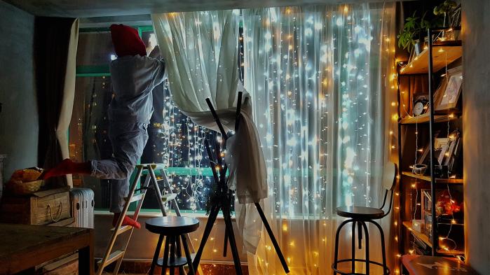 Man with cap on head hangs string of lights on his window while standing on ladder