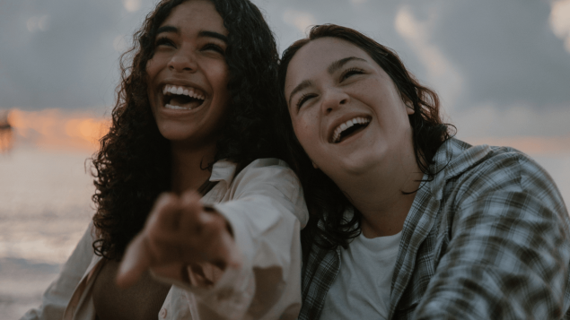 Zwei junge Frauen lehnen sich aneinander und lachen.