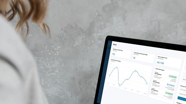 Eine Person betrachtet ein Laptop-Display mit einem Dashboard, das Diagramme und Statistiken zeigt, vor einer grauen Wand.