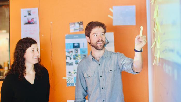 Man and woman standing at whiteboard smiling with post-it in hand