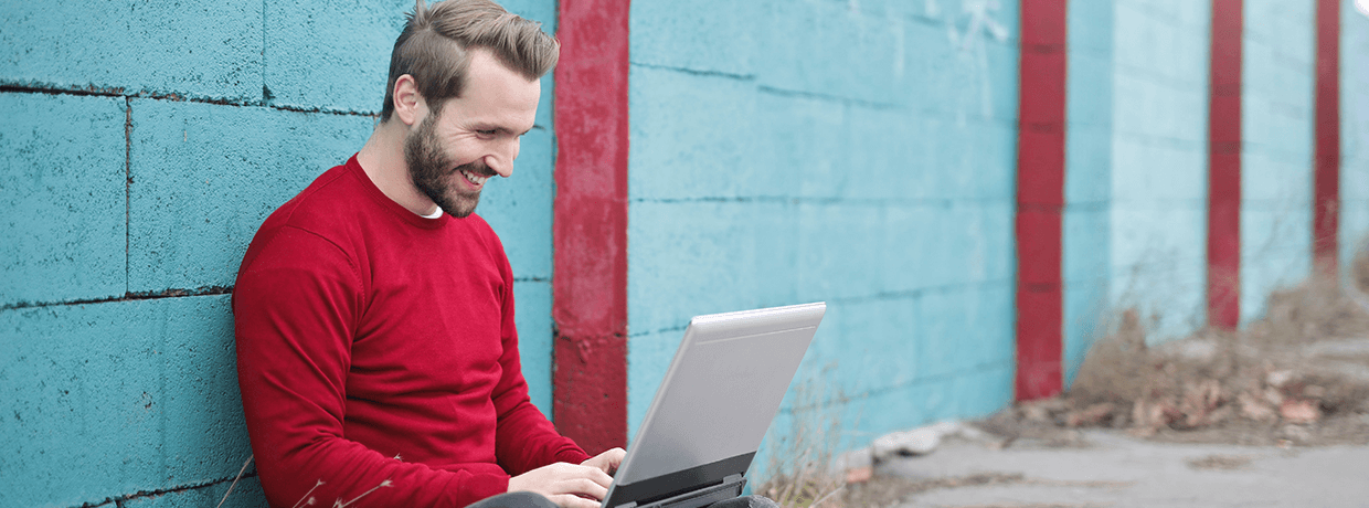 Ein Mann sitzt neben einer blauen Wand und schaut glücklich auf einen Laptop.