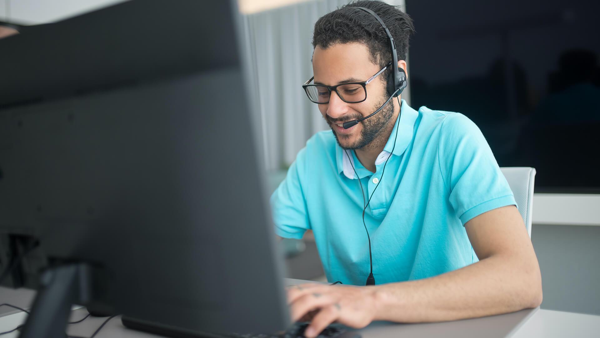 Man with turquoise shirt typing on his computer while talking on headset on phone
